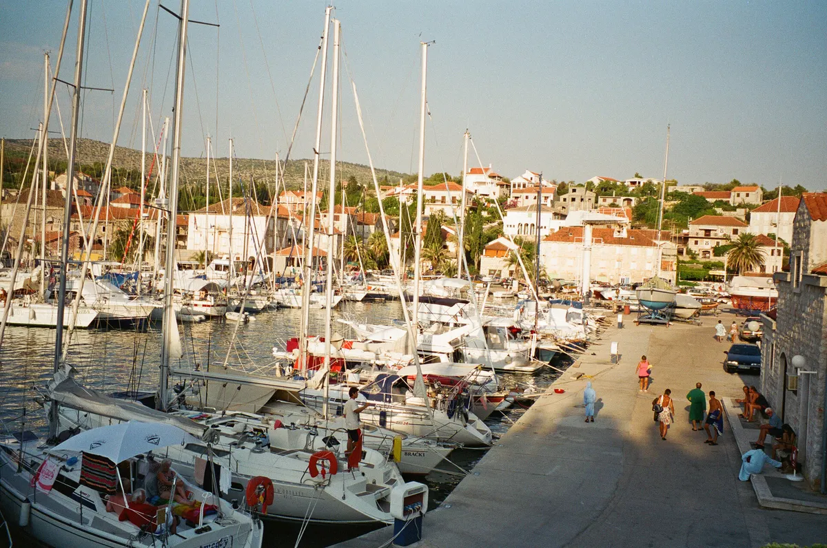 ACI Marina Milna from above the quay: yachts moored stern-to along the marina's pontoons and quay, people walking on the quay, boats on the hardstanding at its end, and the stone houses of Milna on the slope behind