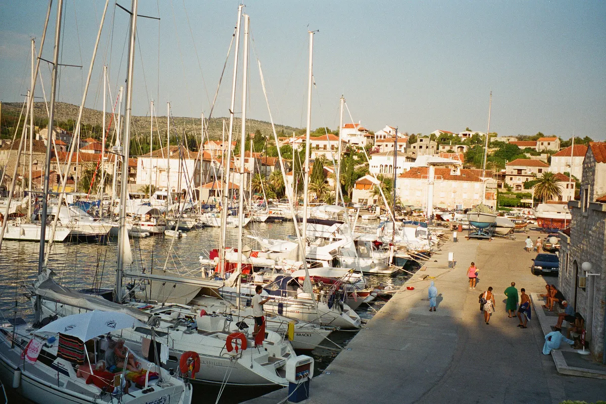 ACI Marina Milna from above the quay: yachts moored stern-to along the marina's pontoons and quay, people walking on the quay, boats on the hardstanding at its end, and the stone houses of Milna on the slope behind
