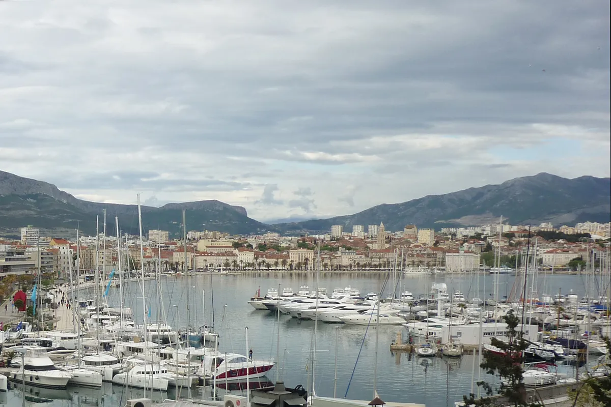ACI Marina Split from Sustipan park: motor boats and yachts on the pontoons in the foreground, the Split waterfront and old town across the harbour, and the Kozjak and Mosor mountains behind under cloud