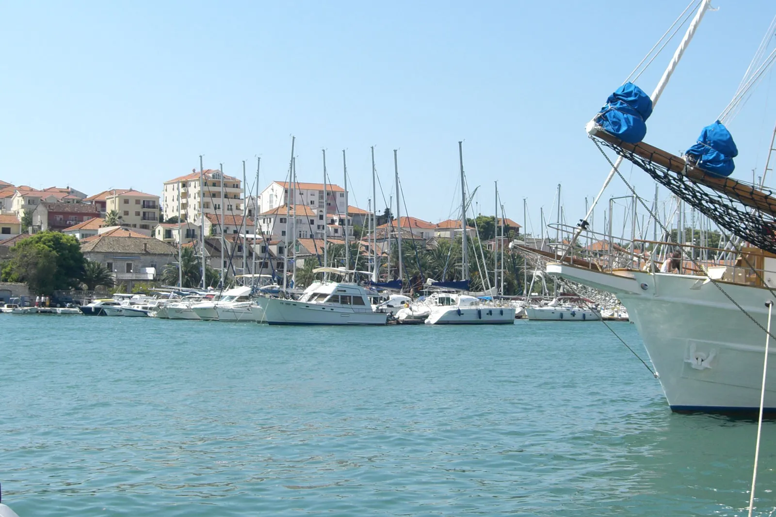 ACI Marina Trogir on the Čiovo shore seen across the channel from the Trogir Riva, yachts and motor boats along the pontoons, the bow of a moored tall ship at the right