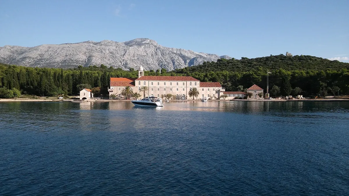 The Franciscan monastery on Badija seen from the water, its quay with a motorboat, pine woods behind and the mountains of Pelješac beyond