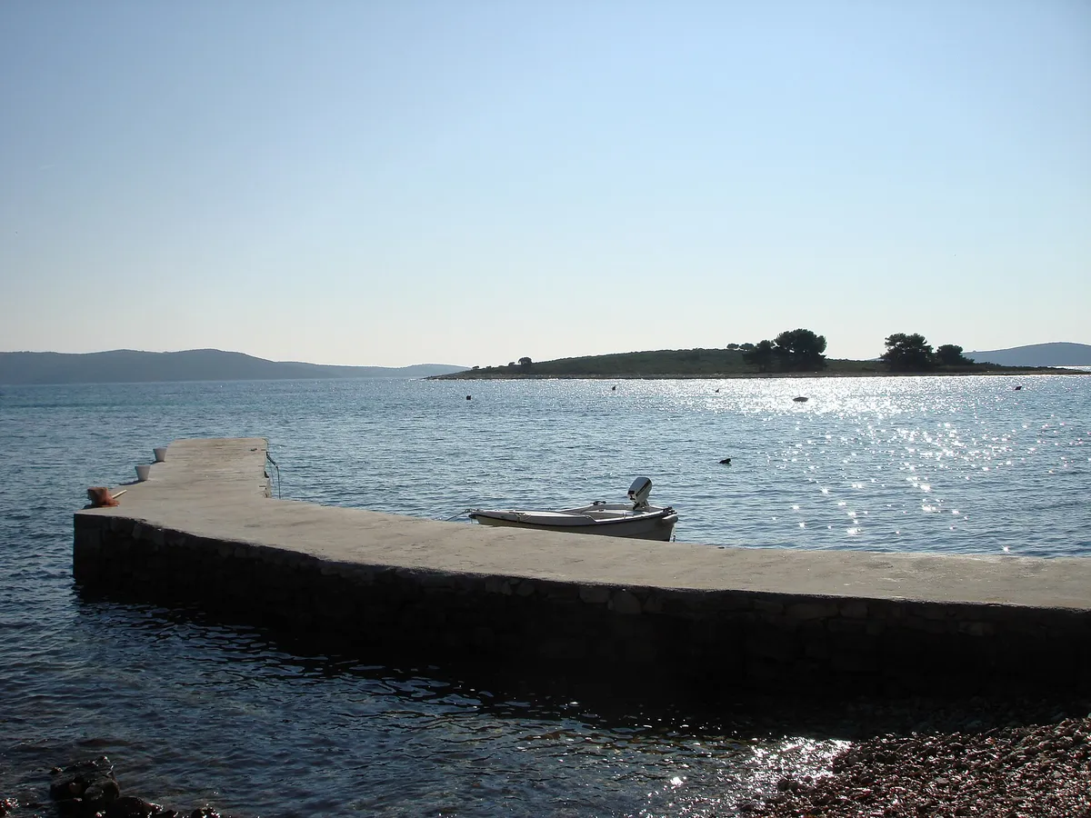 The islet of Veli Krknjaš seen across the calm water of the Blue Lagoon from a small pier at Krknjaši, with a dinghy tied alongside