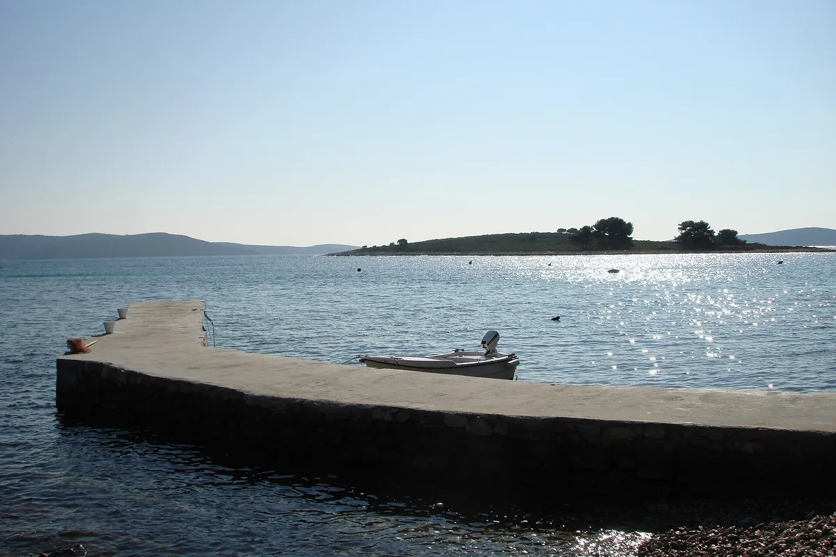 The islet of Veli Krknjaš seen across the calm water of the Blue Lagoon from a small pier at Krknjaši, with a dinghy tied alongside