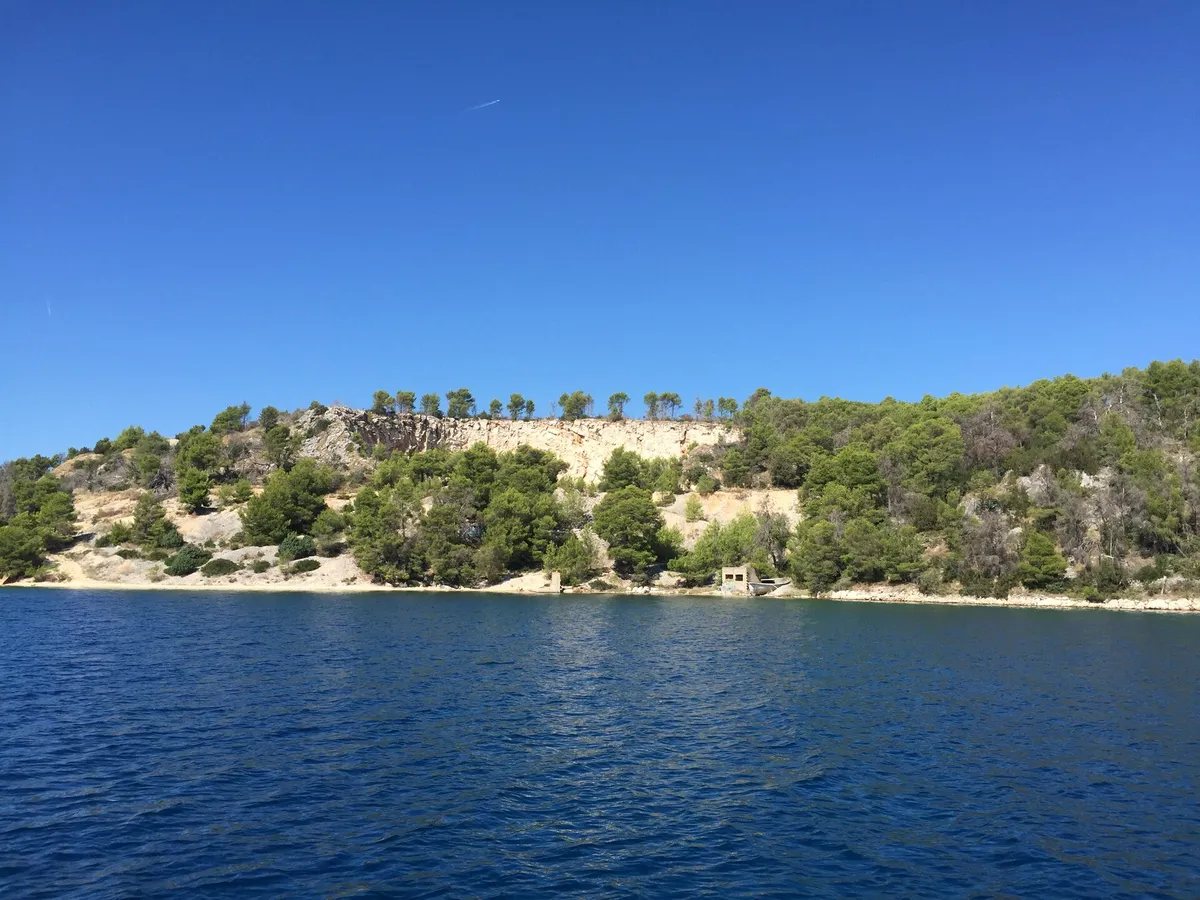 A shore of the outer bay at Bobovišća on Brač, seen from the water where the buoys lie: a pale old quarry face, pines and a small stone hut by the sea