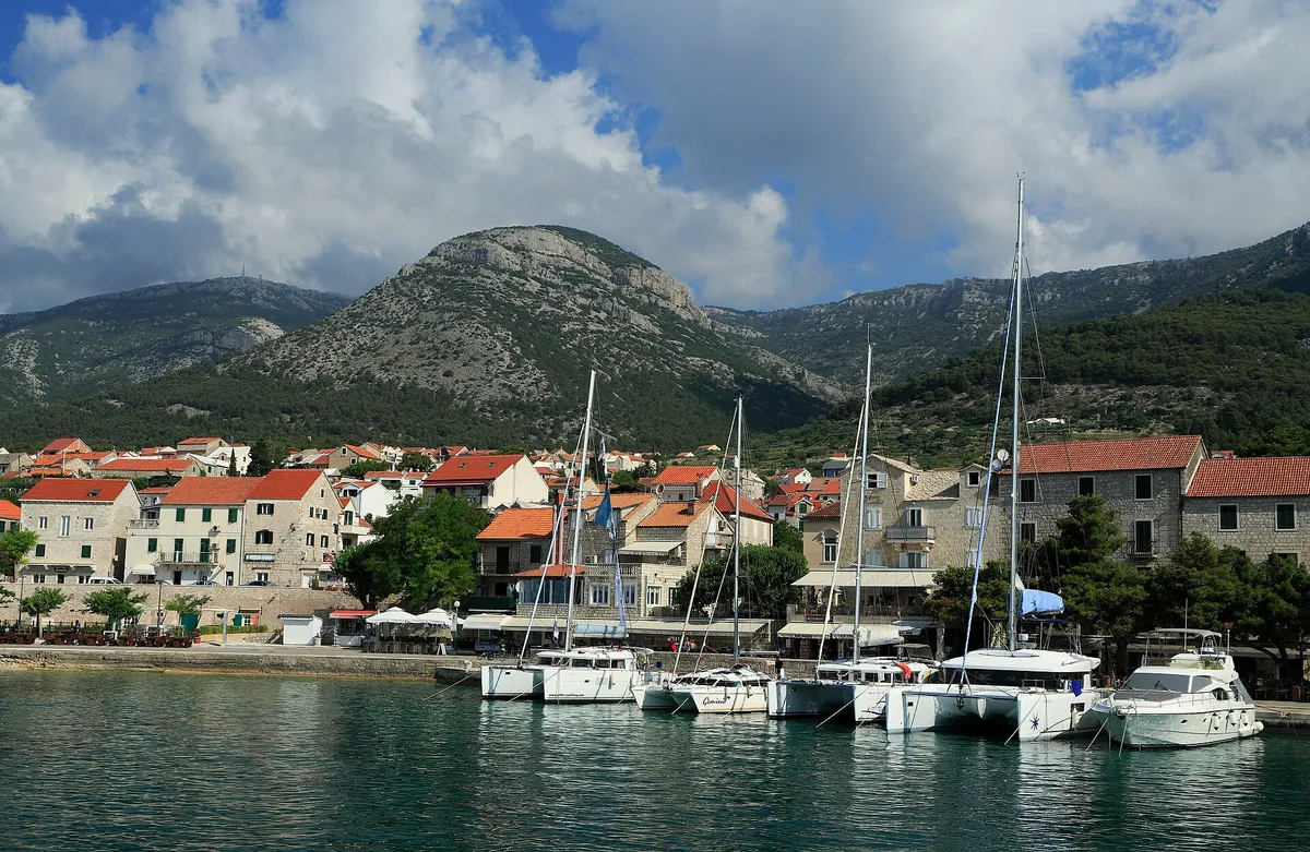Charter catamarans and yachts moored stern-to on the town quay at Bol, seen from a boat leaving the harbour, with Vidova Gora and Velike Koštilo behind.