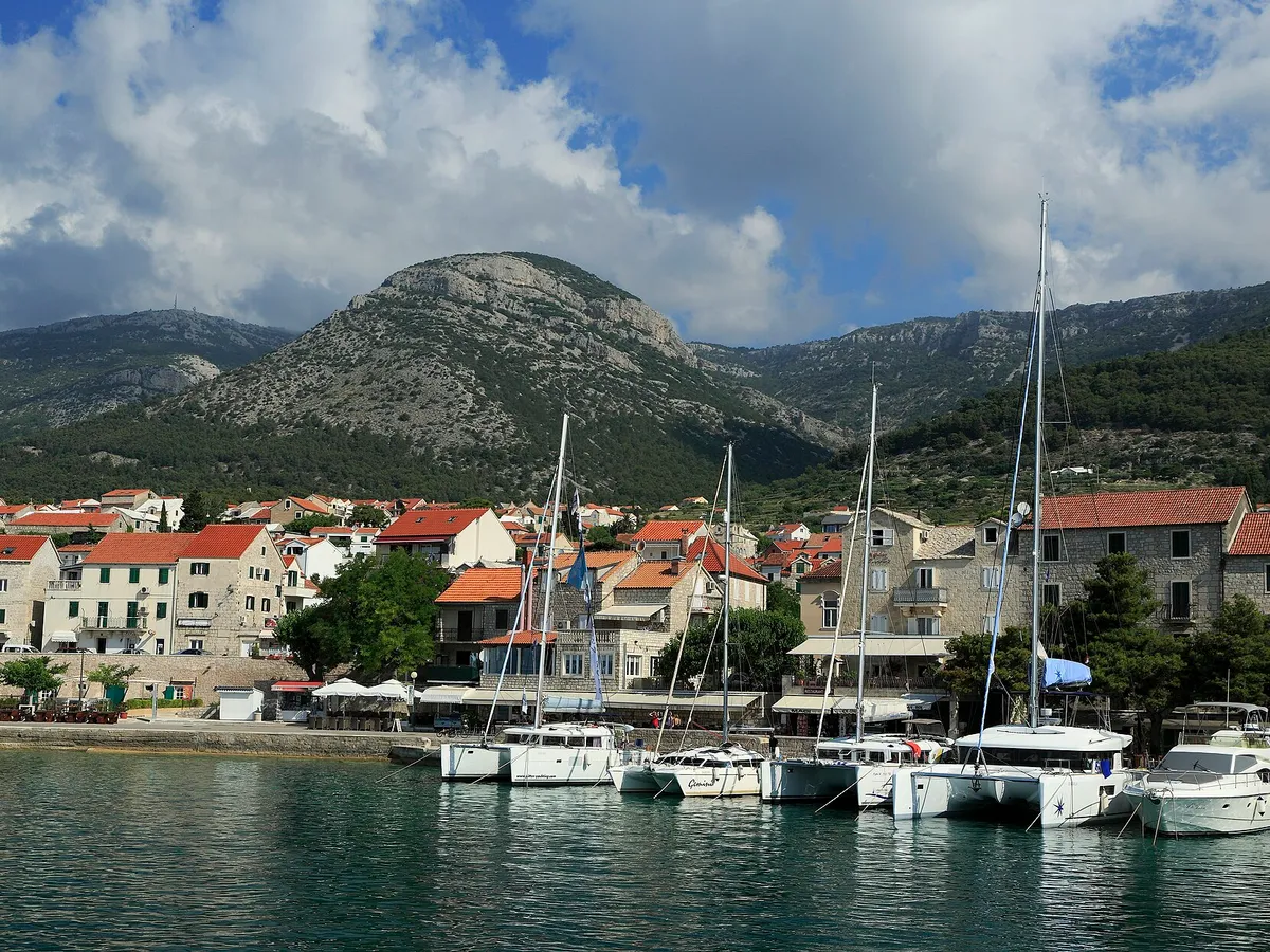 Charter catamarans and yachts moored stern-to on the town quay at Bol, seen from a boat leaving the harbour, with Vidova Gora and Velike Koštilo behind.
