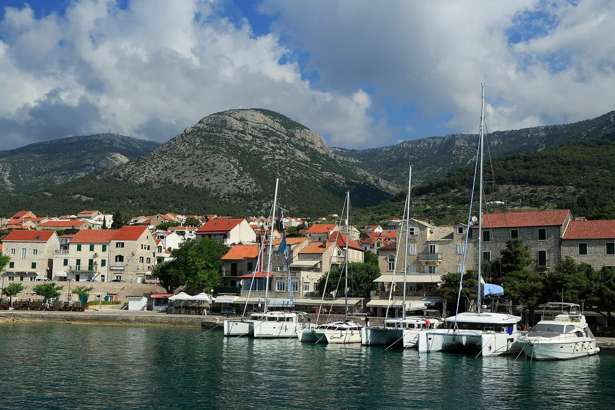 Charter catamarans and yachts moored stern-to on the town quay at Bol, seen from a boat leaving the harbour, with Vidova Gora and Velike Koštilo behind.
