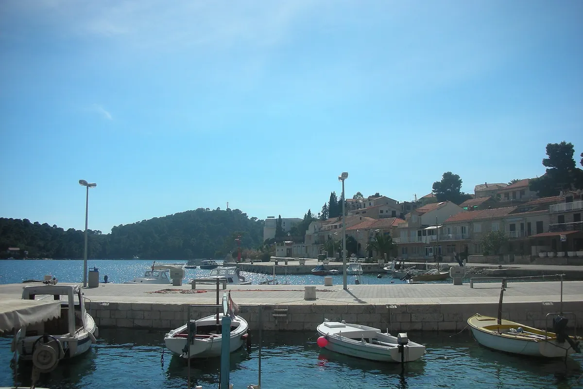 The harbour at Brna on Korčula, with small wooden and motor boats at a stone quay, a paved waterfront and houses along the shore, and a wooded headland across the bay