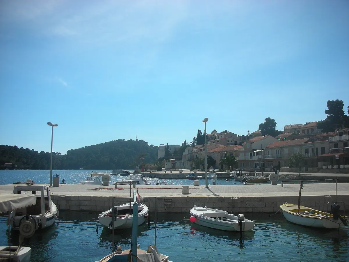 The harbour at Brna on Korčula, with small wooden and motor boats at a stone quay, a paved waterfront and houses along the shore, and a wooded headland across the bay
