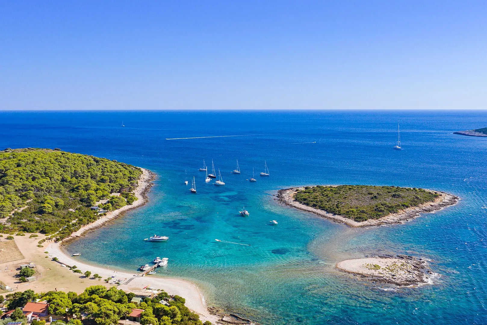 The lagoon between Veli Budikovac and Mali Budikovac from the air, yachts on the buoys in turquoise water, the pebble beach and the jetty on Veli Budikovac and open sea beyond
