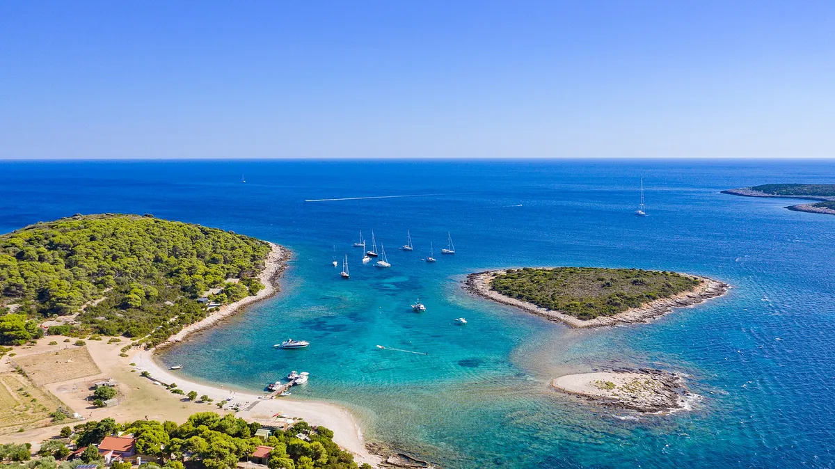 The lagoon between Veli Budikovac and Mali Budikovac from the air, yachts on the buoys in turquoise water, the pebble beach and the jetty on Veli Budikovac and open sea beyond