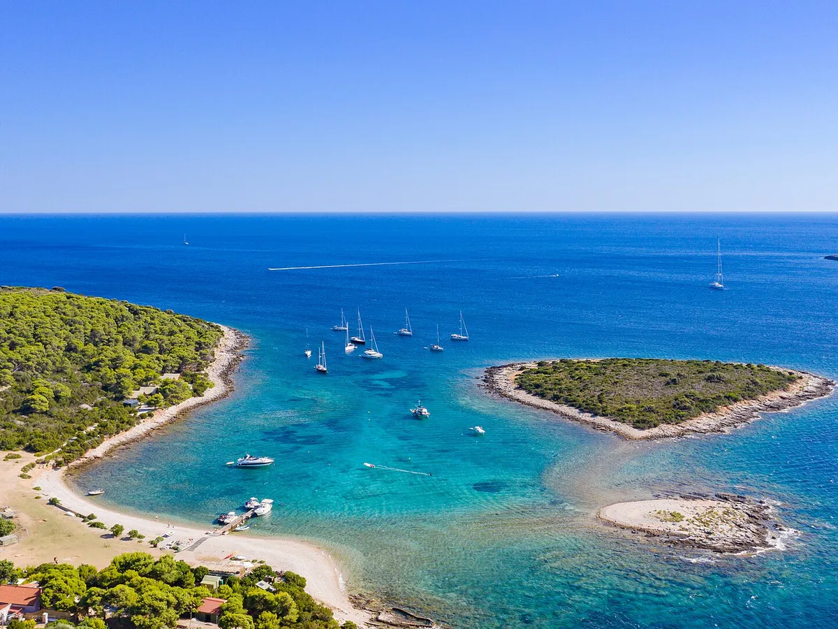 The lagoon between Veli Budikovac and Mali Budikovac from the air, yachts on the buoys in turquoise water, the pebble beach and the jetty on Veli Budikovac and open sea beyond