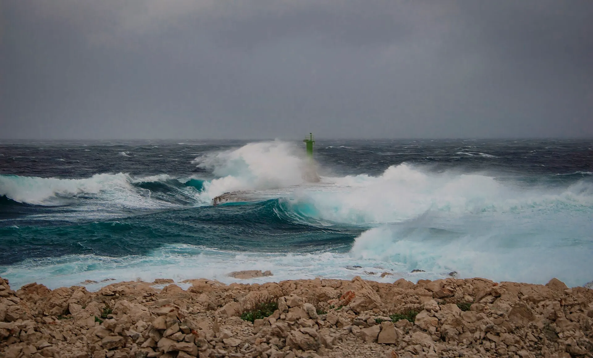 A storm on the Adriatic with waves breaking over a harbour wall and its green light, under a grey sky.