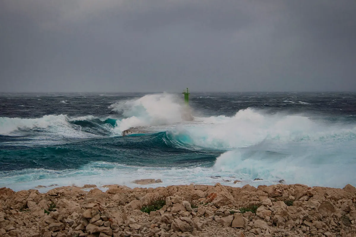A storm on the Adriatic with waves breaking over a harbour wall and its green light, under a grey sky.