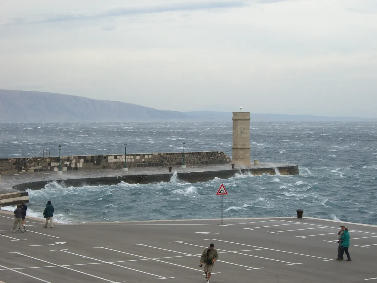 A bura day in Senj: the harbour breakwater and its light in a sea whipped white by the wind, with people on the quay leaning into it and the islands beyond