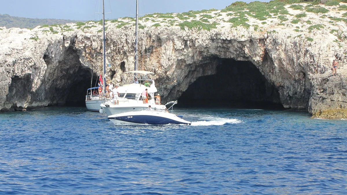 The two sea entrances of the Green Cave on Ravnik seen from the water, a sailing yacht and a motor cruiser at the mouth and a speedboat passing