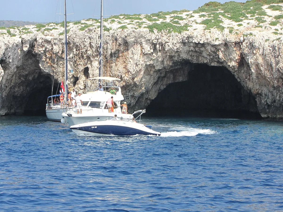 The two sea entrances of the Green Cave on Ravnik seen from the water, a sailing yacht and a motor cruiser at the mouth and a speedboat passing
