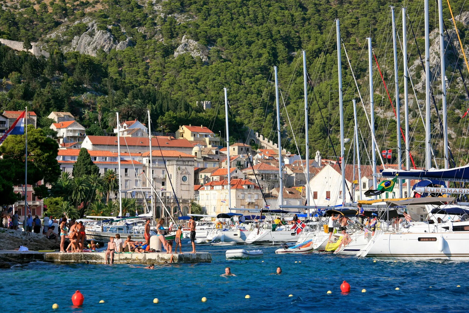 Yachts moored side by side along the quay in Hvar town, with old stone houses, the clock tower and the pine-covered hill behind, and swimmers on a stone bathing platform and in the water in front