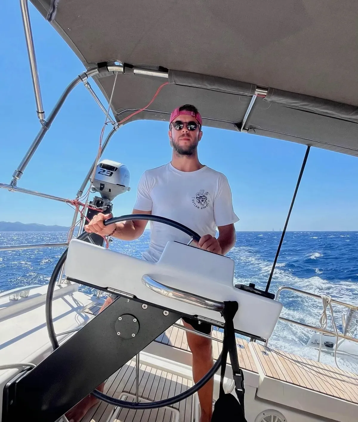 Ivor Benko at the helm of a sailing yacht under way, under the bimini, with a blue sea and the coast behind