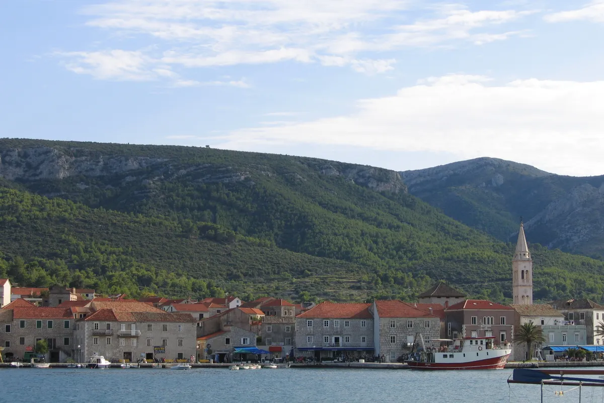 Jelsa on Hvar across its harbour: the stone waterfront with the church tower, boats at the quay and pine-covered hills behind