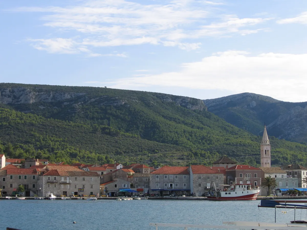 Jelsa on Hvar across its harbour: the stone waterfront with the church tower, boats at the quay and pine-covered hills behind
