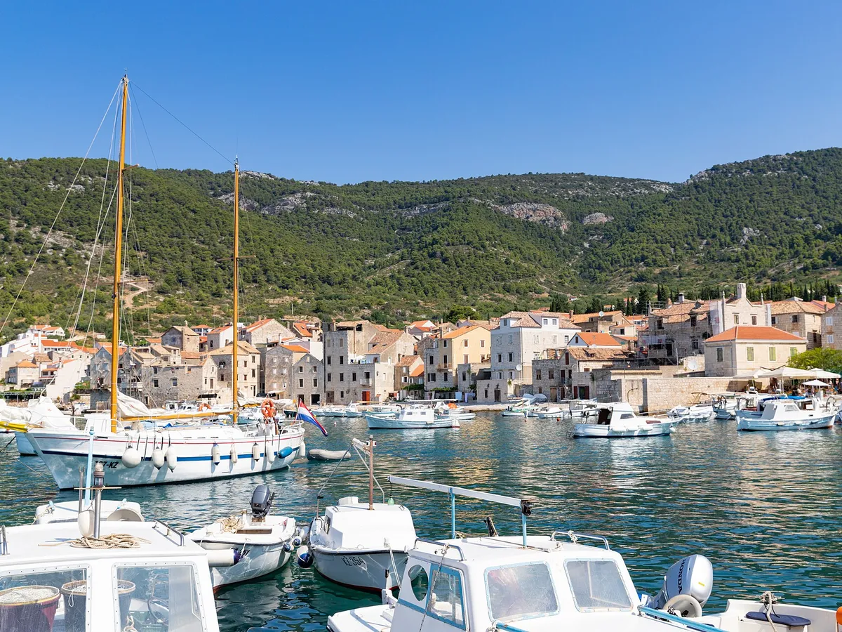 Komiža harbour on Vis, with fishing boats, a wooden sailing boat and small motorboats in front of the old town and the hills.