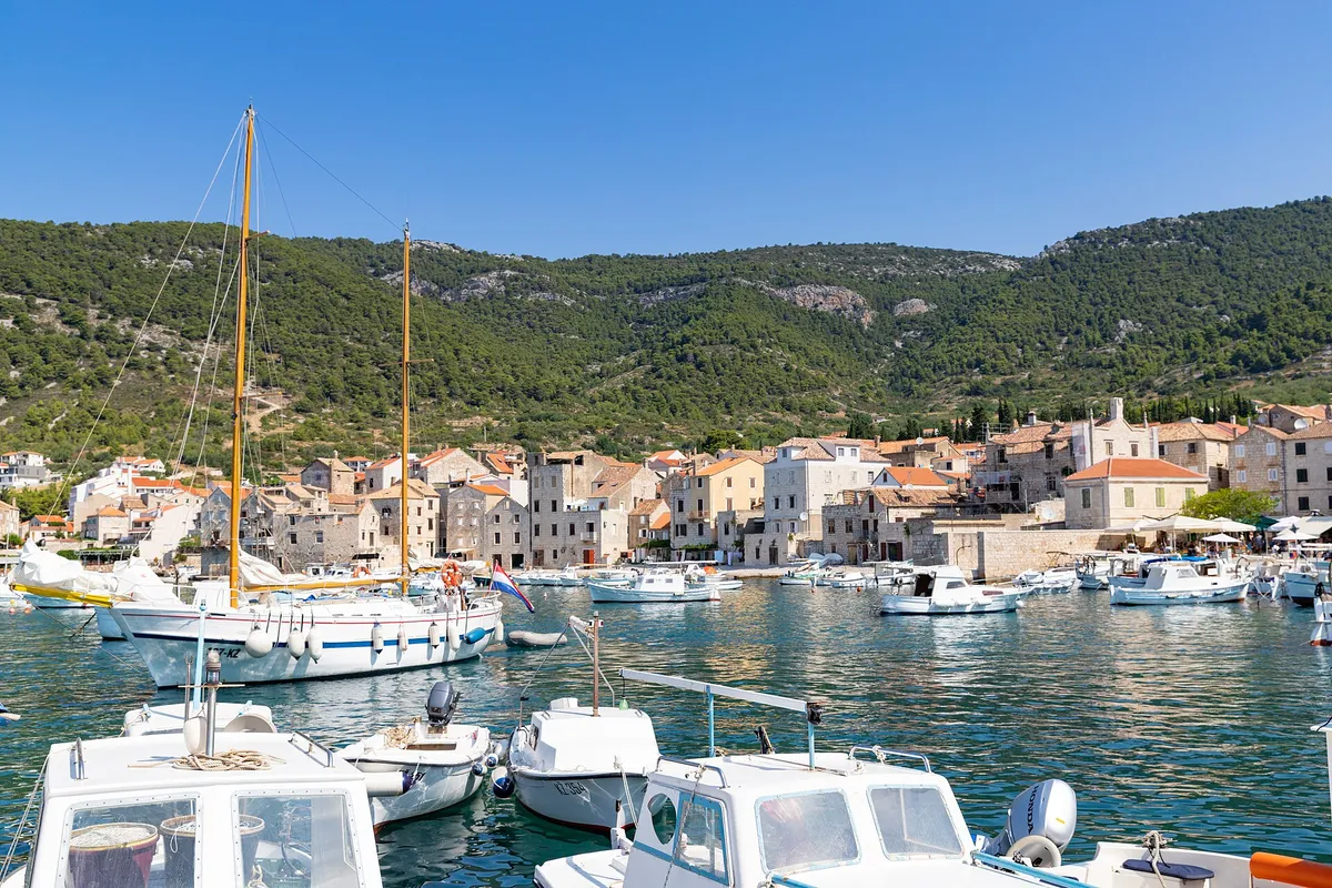 Komiža harbour on Vis, with fishing boats, a wooden sailing boat and small motorboats in front of the old town and the hills.