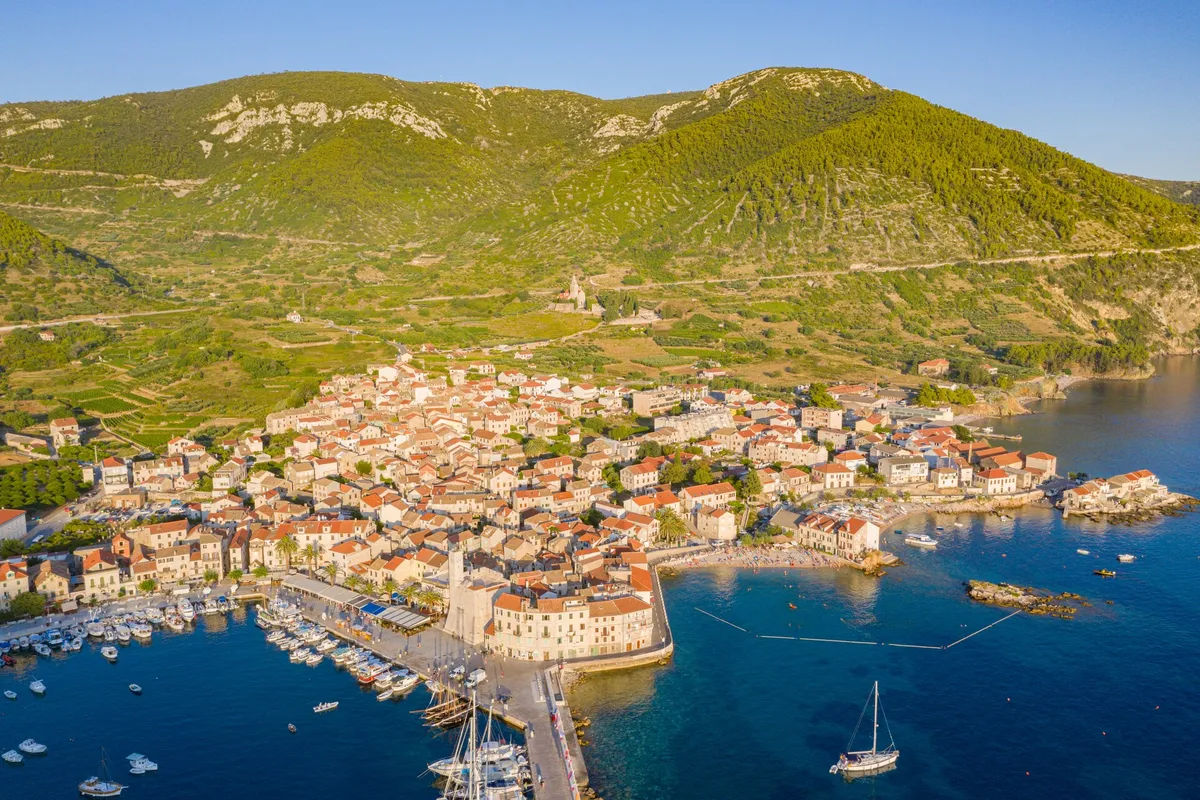 Komiža from the air on a summer evening, the harbour and its pier on the left and, right of the Kaštel, the cove of Lučica beach with its rocky islet, swimmers and a sailing yacht