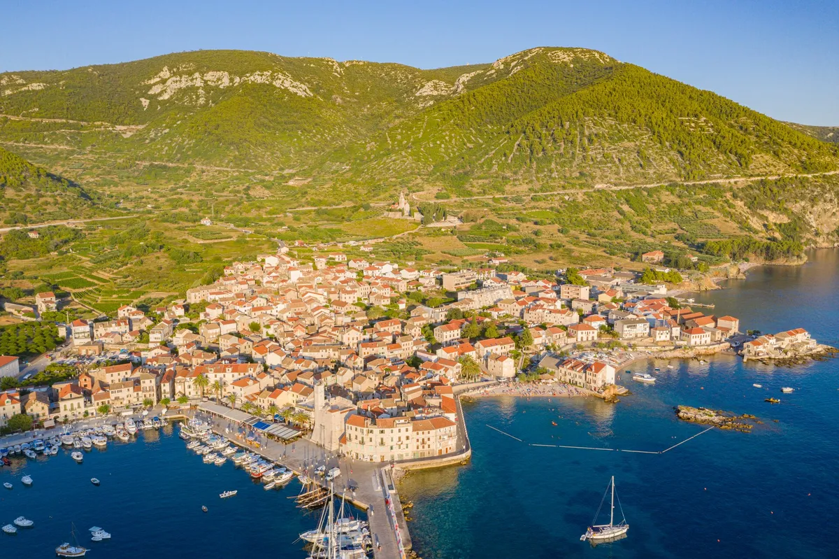 Komiža from the air on a summer evening, the harbour and its pier on the left and, right of the Kaštel, the cove of Lučica beach with its rocky islet, swimmers and a sailing yacht