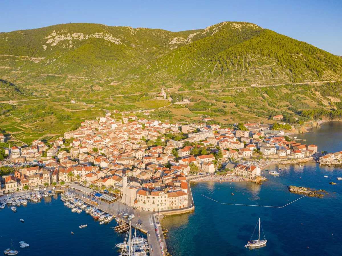 Komiža from the air on a summer evening, the harbour and its pier on the left and, right of the Kaštel, the cove of Lučica beach with its rocky islet, swimmers and a sailing yacht