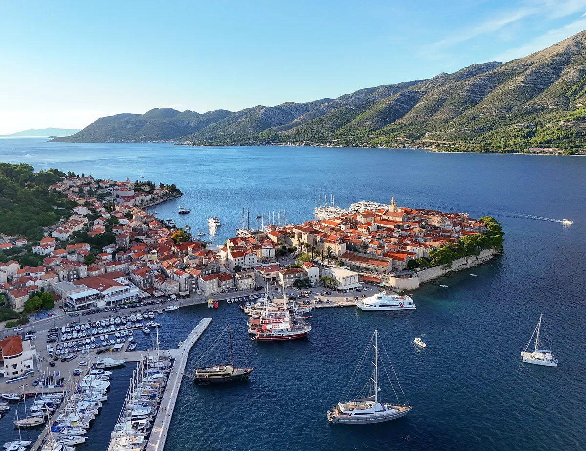Korčula old town on its peninsula seen from the air, with ACI Marina Korčula and the east harbour in front, sailing yachts and gulets at the quays, and the Pelješac mountains across the channel