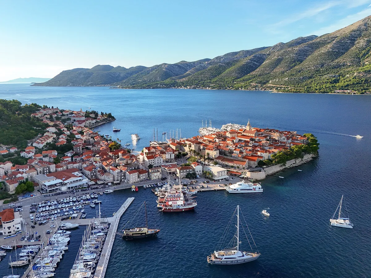 Korčula old town on its peninsula seen from the air, with ACI Marina Korčula and the east harbour in front, sailing yachts and gulets at the quays, and the Pelješac mountains across the channel
