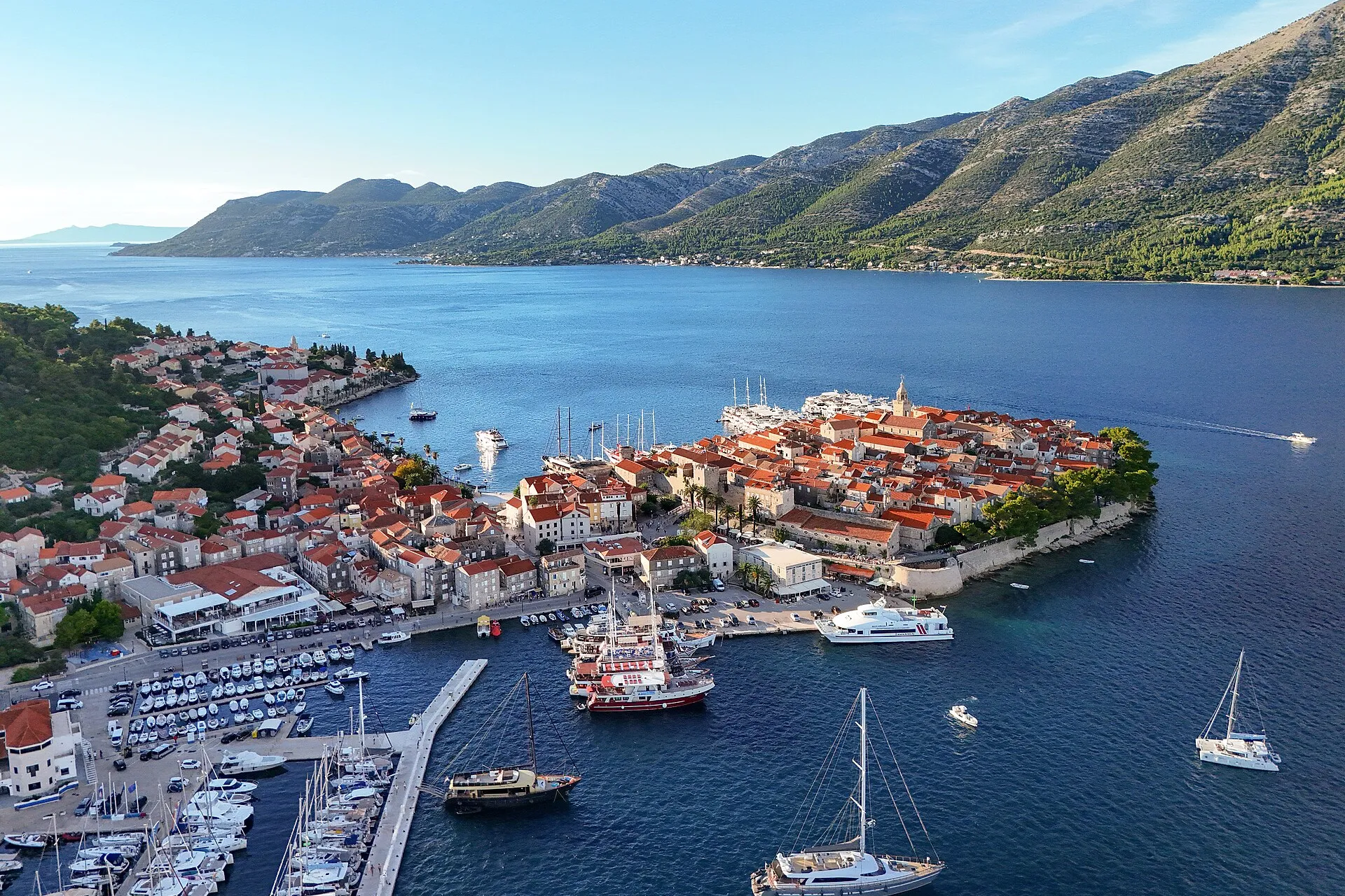 Korčula old town on its peninsula seen from the air, with ACI Marina Korčula and the east harbour in front, sailing yachts and gulets at the quays, and the Pelješac mountains across the channel