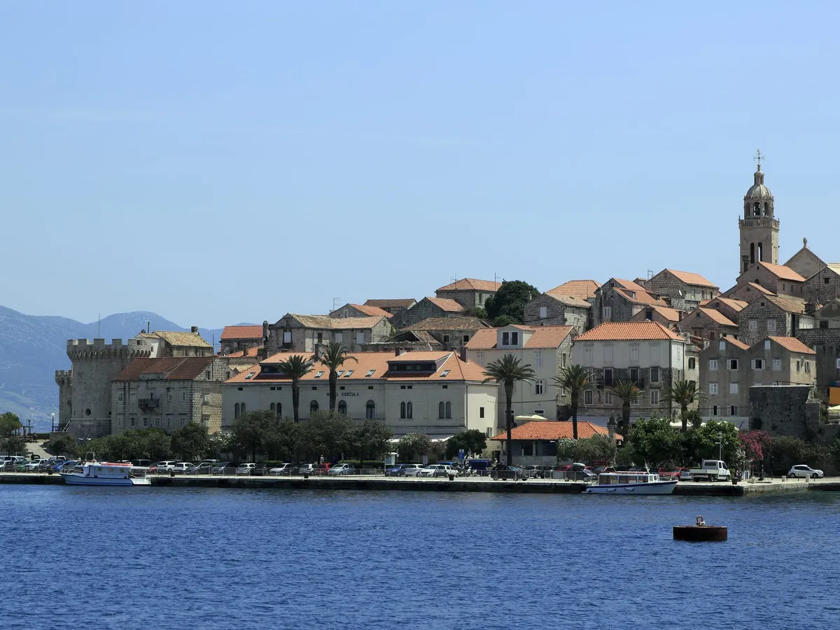 The old town of Korčula from the water, with stone houses, a round tower and the bell tower above the town quay, where boats are moored