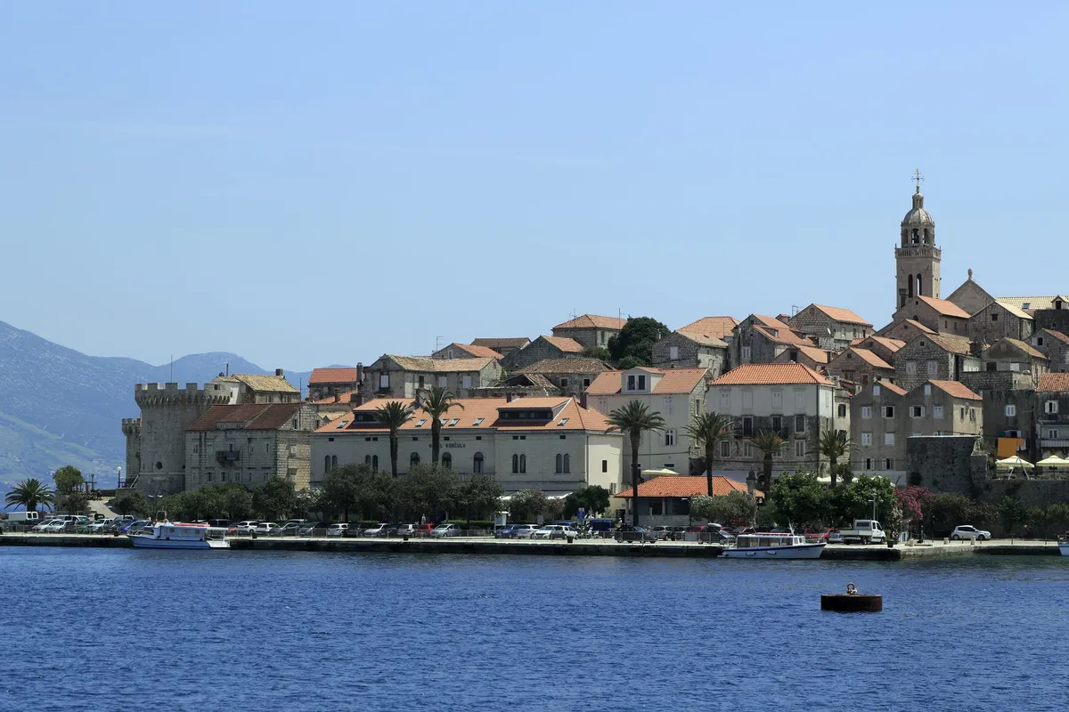 The old town of Korčula from the water, with stone houses, a round tower and the bell tower above the town quay, where boats are moored