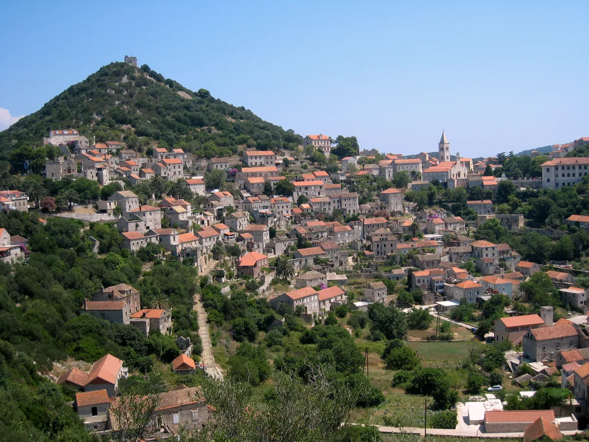 Lastovo town on its hillside, stone houses with red roofs climbing towards the church tower, below the wooded hill with the fort on top