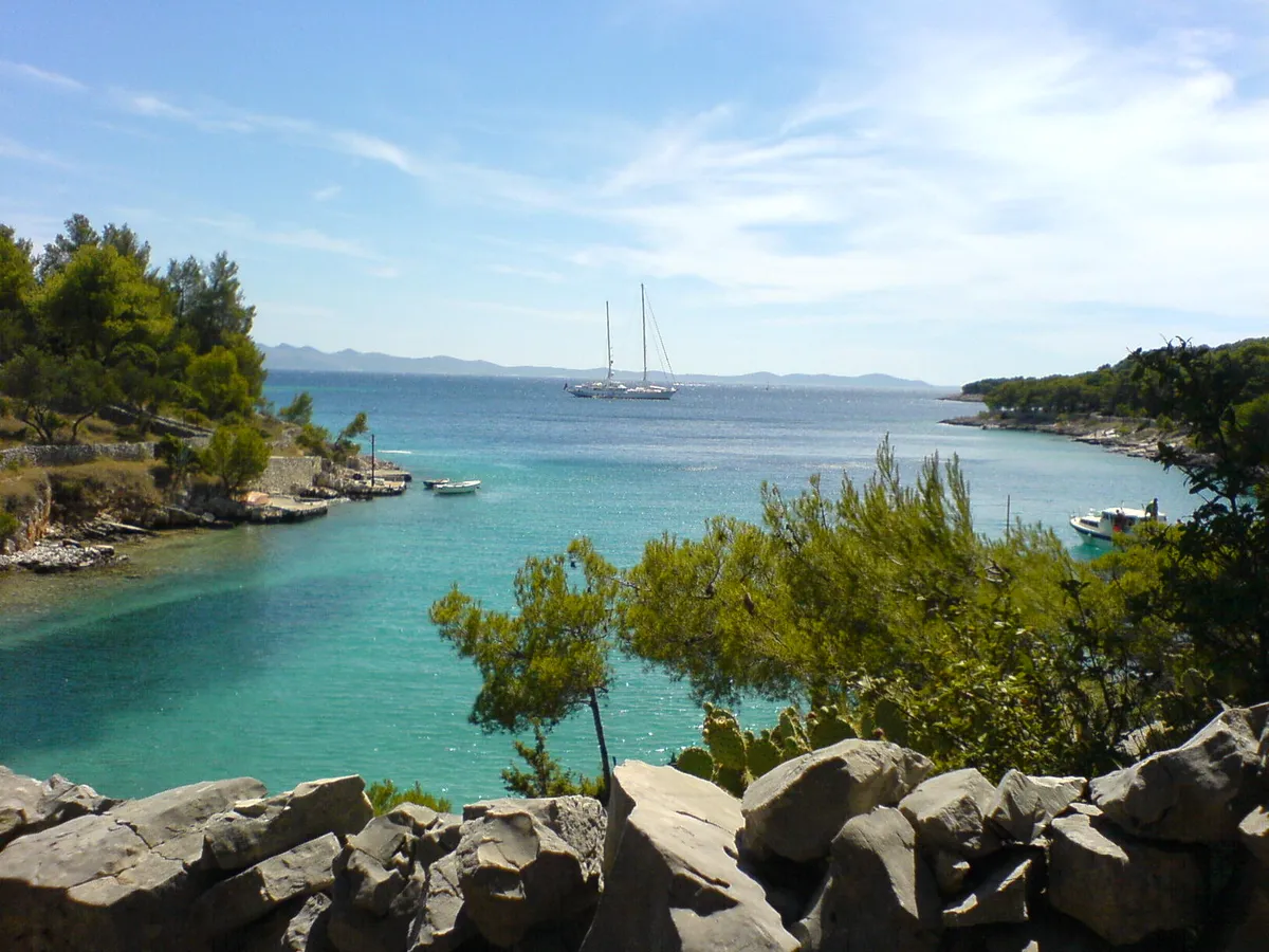 Lučice bay seen from the rocks above it, turquoise water between pine-covered arms, a two-masted yacht at anchor at the mouth and small boats by the shore