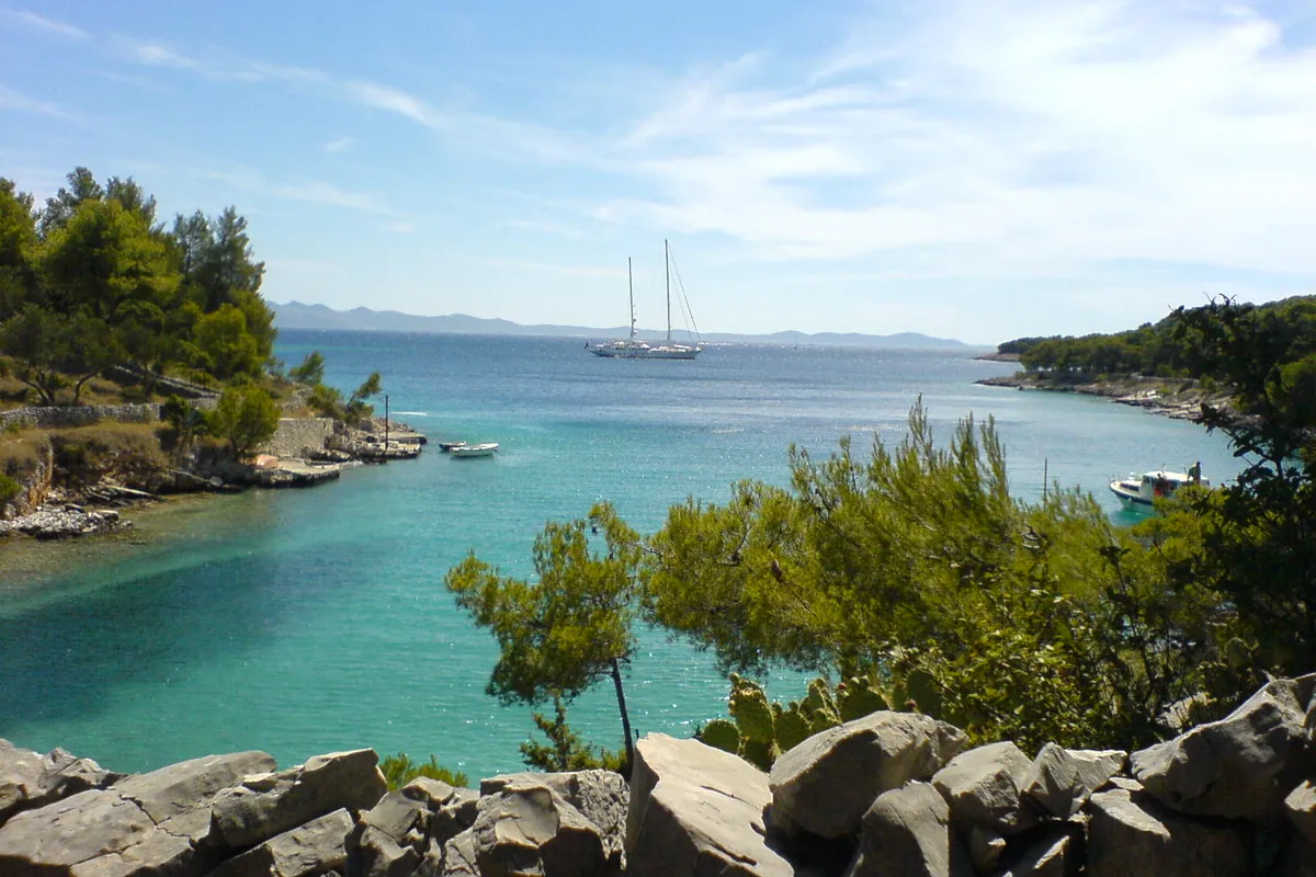 Lučice bay seen from the rocks above it, turquoise water between pine-covered arms, a two-masted yacht at anchor at the mouth and small boats by the shore