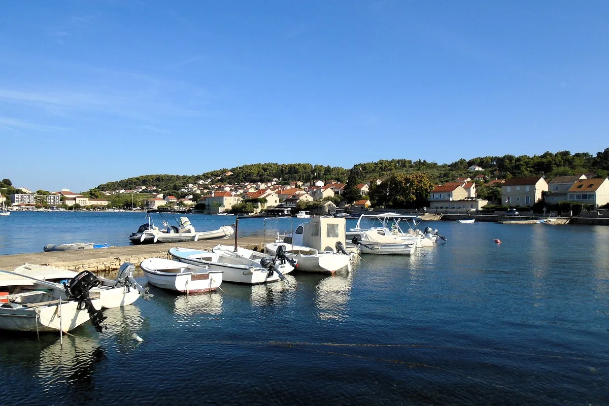 Lumbarda's harbour on a calm morning, with small motor boats tied to a stone pier, houses and pine woods on the hill behind, and a few yacht masts across the bay