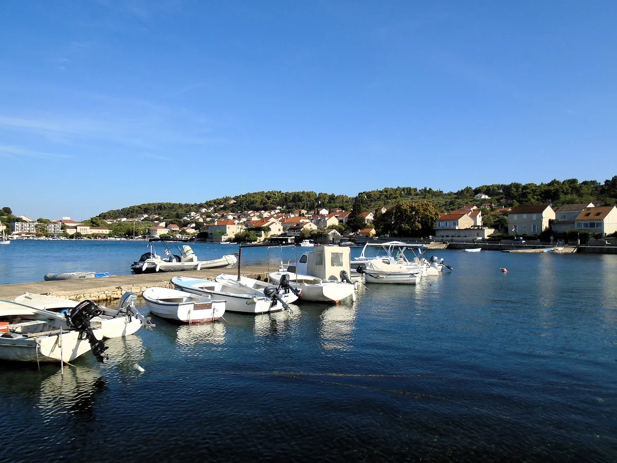 Lumbarda's harbour on a calm morning, with small motor boats tied to a stone pier, houses and pine woods on the hill behind, and a few yacht masts across the bay