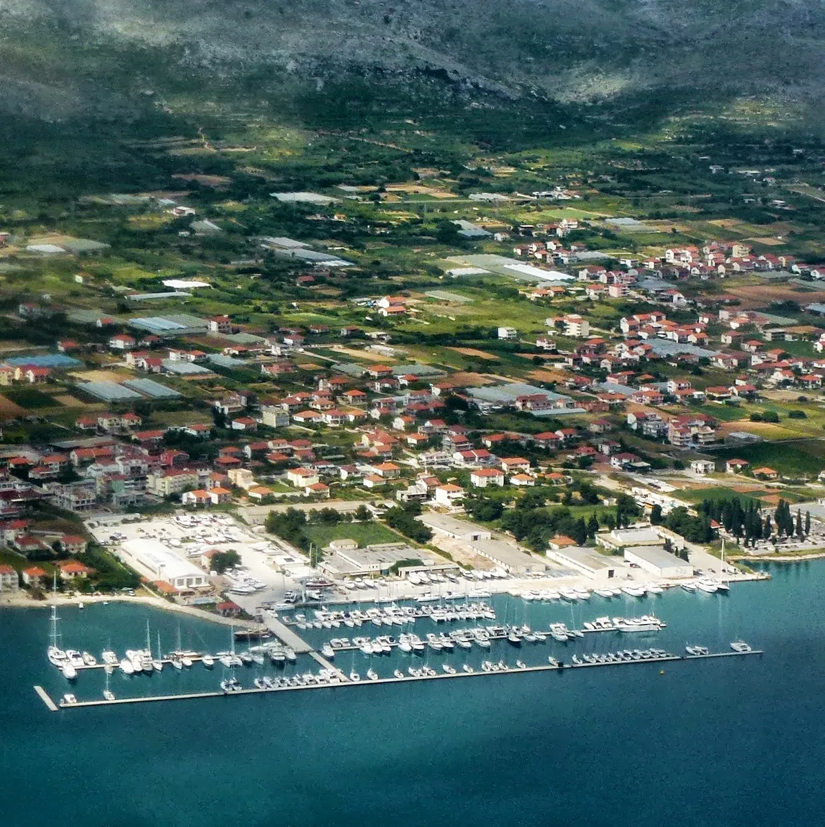 Marina Baotić at Seget Donji from the air: long pontoons full of yachts off the shore, the boatyard behind them and the village among fields