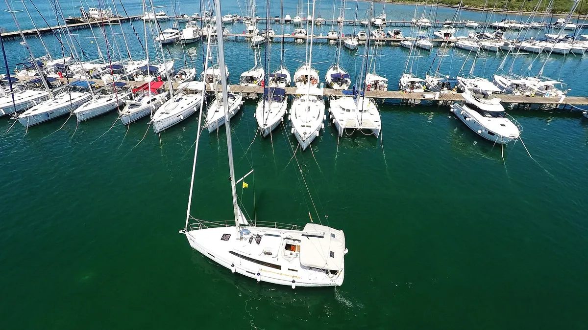 A drone view of yachts moored stern-to on a pontoon with lazy lines, and a sailing yacht leaving its berth.