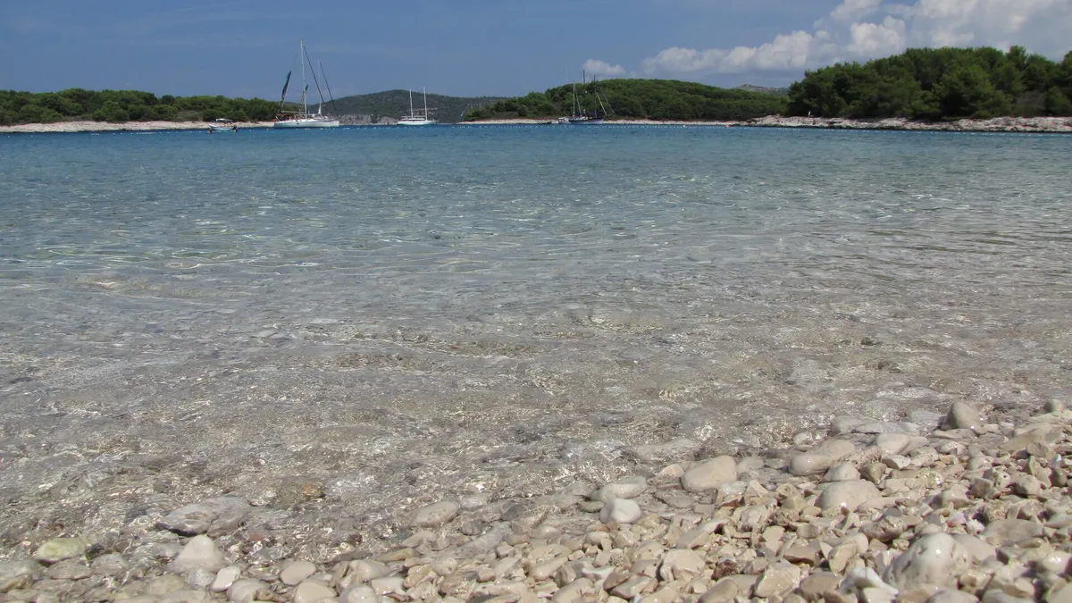 Clear shallow water over the pebbles of Mlini beach on Marinkovac, with yachts at anchor off the low wooded shore across the bay