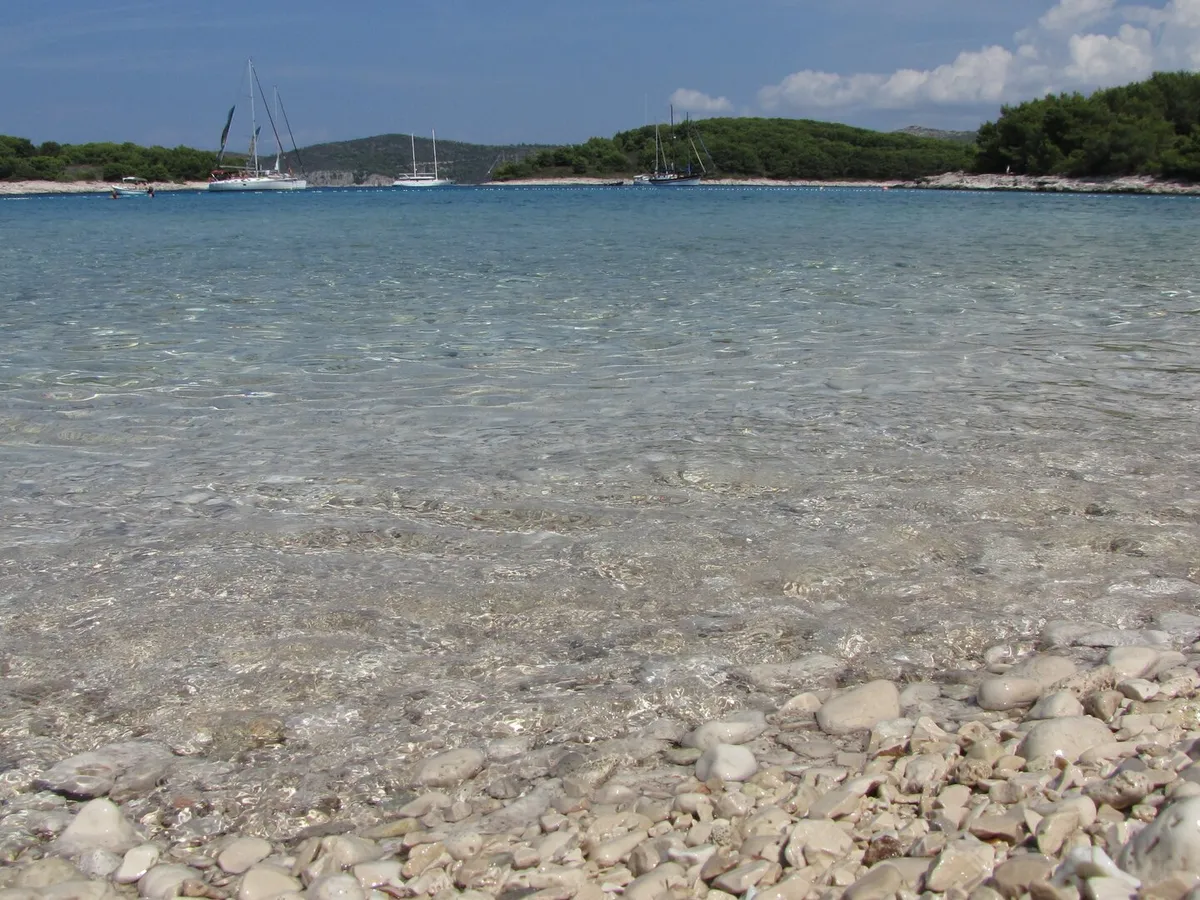 Clear shallow water over the pebbles of Mlini beach on Marinkovac, with yachts at anchor off the low wooded shore across the bay
