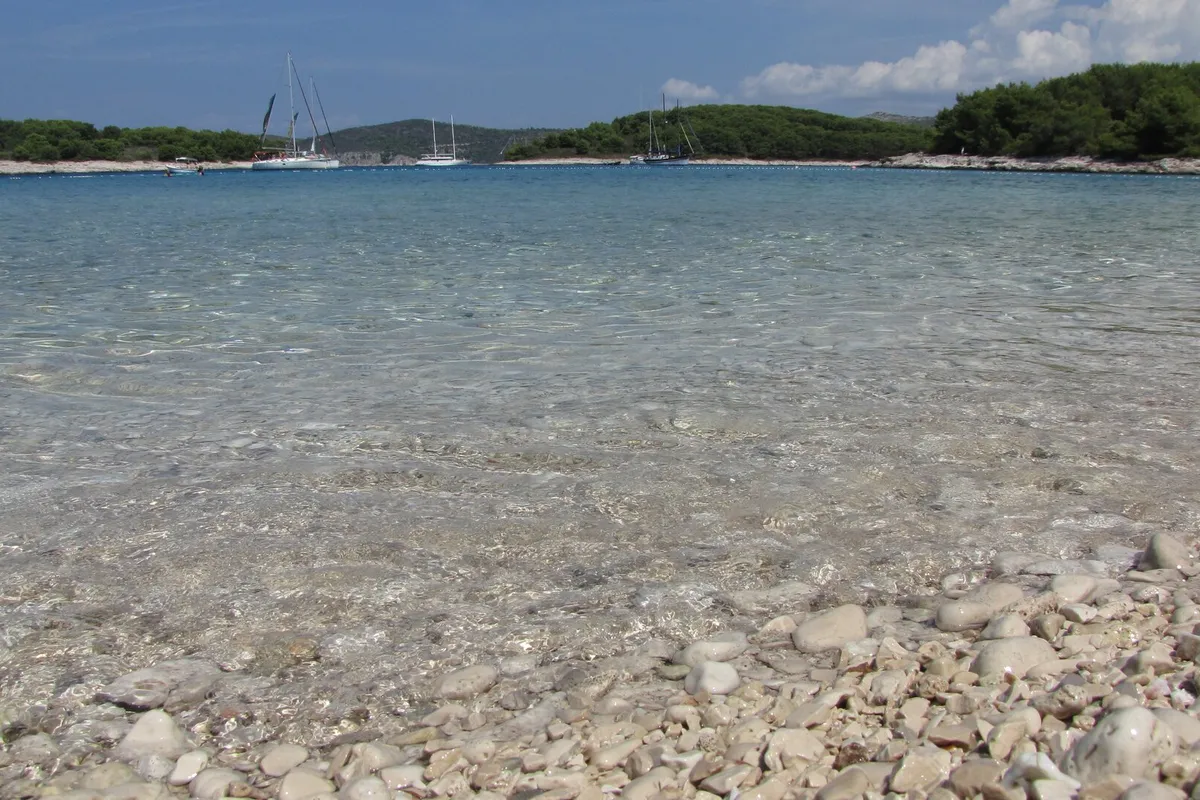 Clear shallow water over the pebbles of Mlini beach on Marinkovac, with yachts at anchor off the low wooded shore across the bay