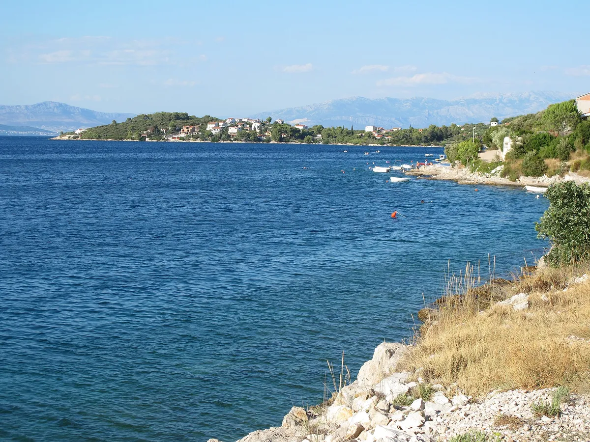 Nečujam bay on Šolta from the rocks on its west shore, looking north-east: small boats along the shore, a mooring buoy in deep blue water and the houses on the far point, with the mountains near Split behind