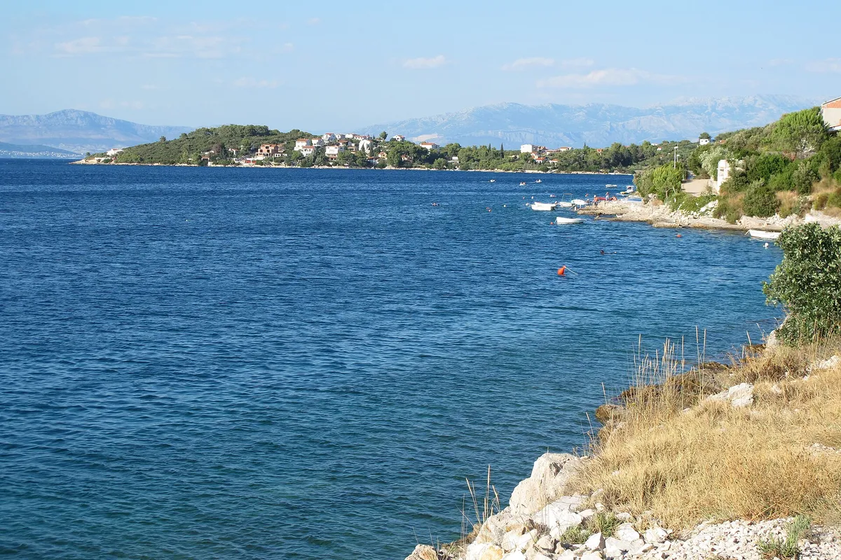 Nečujam bay on Šolta from the rocks on its west shore, looking north-east: small boats along the shore, a mooring buoy in deep blue water and the houses on the far point, with the mountains near Split behind