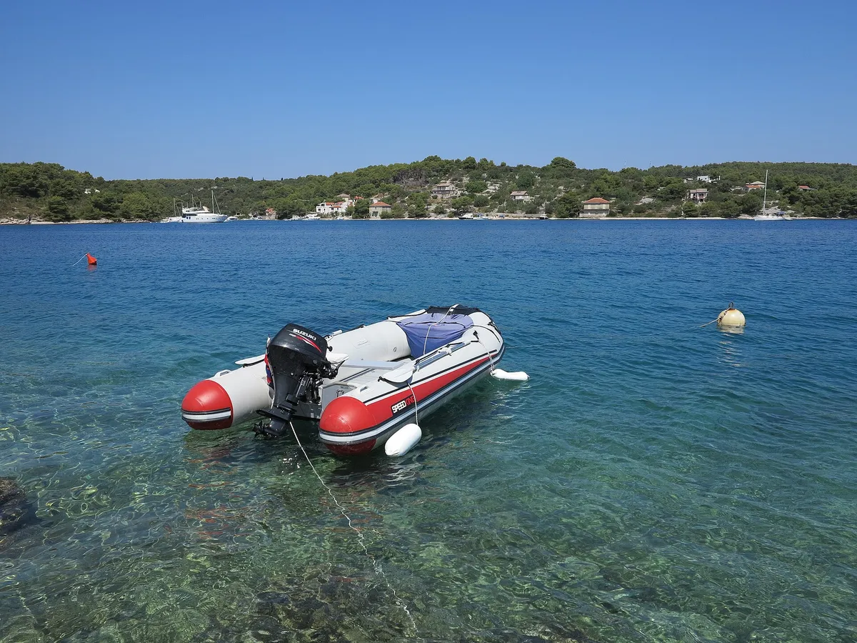 A tender on its mooring over clear water in Podkamenica cove, Nečujam bay, with a mooring buoy beside it and yachts moored along the far shore