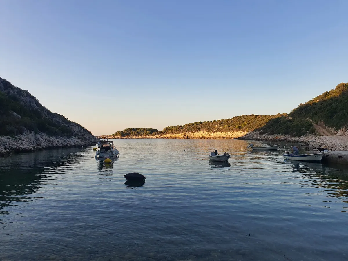 Oključna bay on Vis at dusk, small boats on their moorings in calm water between rocky wooded slopes, the evening sun on the far shore