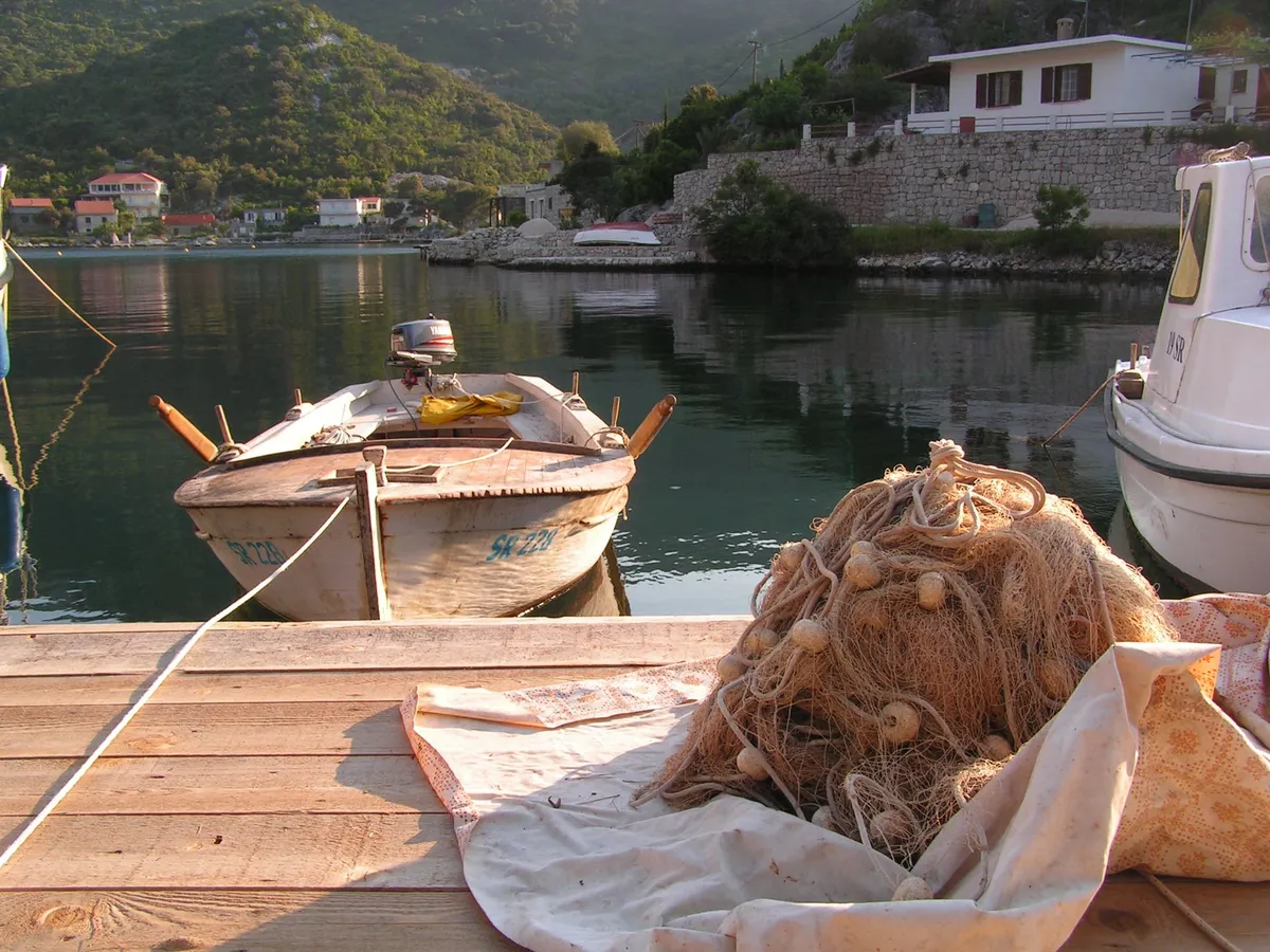 A wooden rowing boat and a fishing net on a jetty in Okuklje bay on Mljet, with the calm bay, the village houses and the wooded hills behind, in evening light